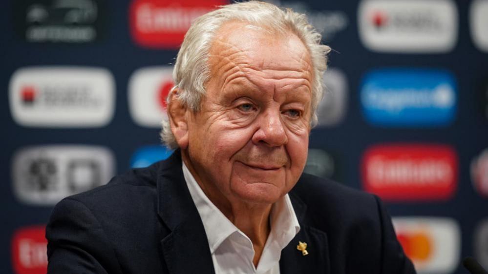 Bill Beaumont with white hair wearing a dark jacket sitting in front of sponsors' signs