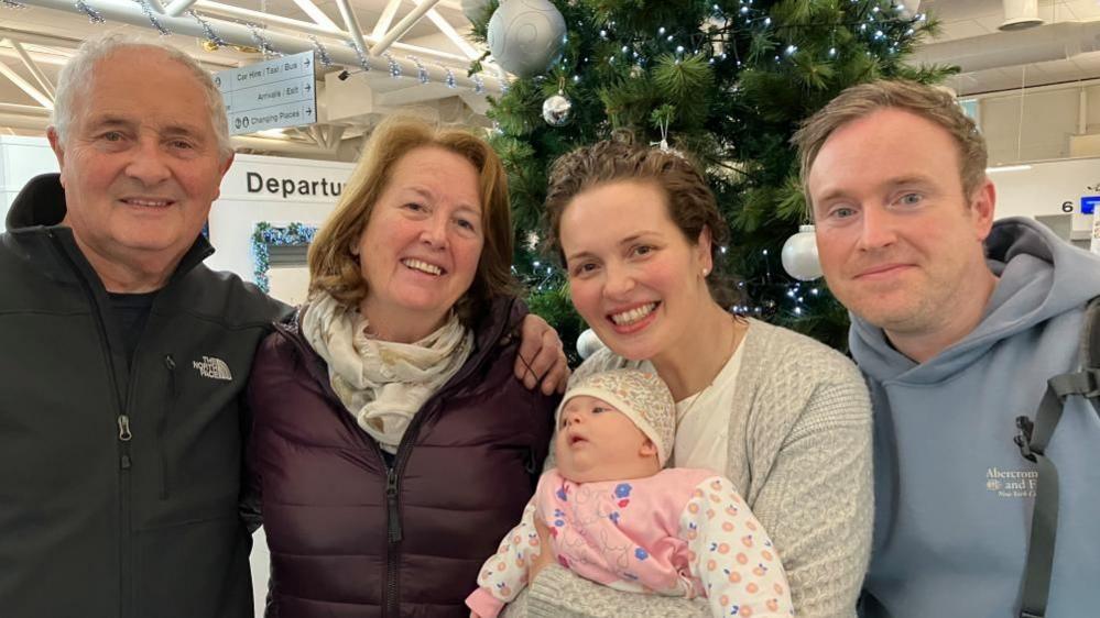 Jack is n the left wearing a black coat, Bernadette is wearing a purple coat and has shoulder length ginger hair. Sinead is holding baby Niamh, who is dressed in pink and wearing a floral hat. Sinead is wearing a beige cardigan. Malachy, right is wearing a blue hoodie and has short ginger hair. 