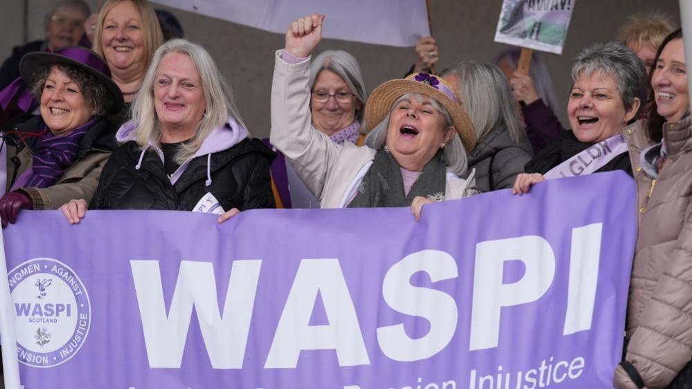 A group of older women wear purple and smile at a demonstration. They are holding signs and a purple banner that reads 'WASPI' 