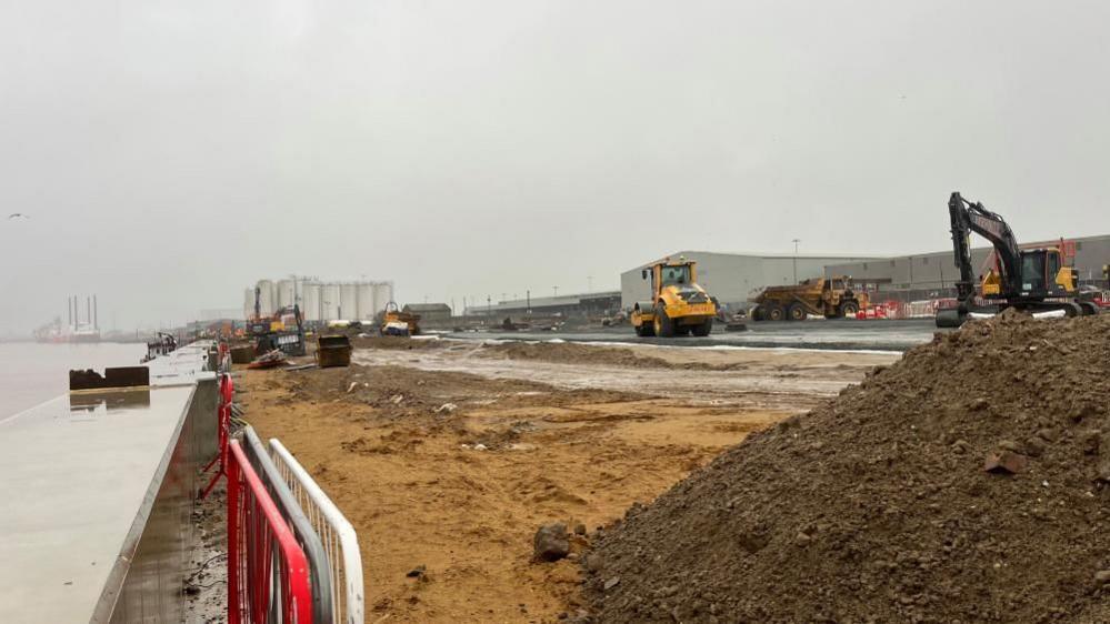 Concrete river wall capping on the left, moving through uneven sand and soil with level ground and earth moving equipment in the background