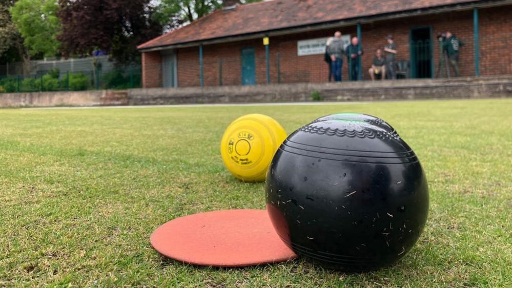 A black bowl, sitting on grass next to a red disc and yellow jack. Several men are sitting outside a brick clubhouse in a blurred background