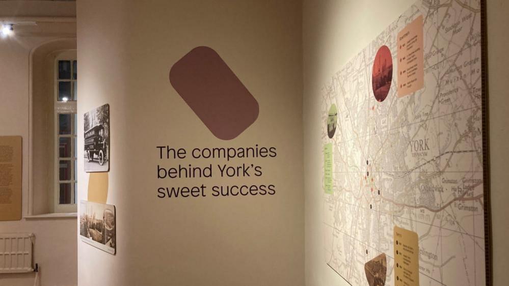 A large display in a room at the Sugar, Skill and Shiftwork: York's Confectionery Workers exhibition at York Castle Museum. In large brown font printed directly onto a white wall reads: The companies behind York's sweet success. To the left, a large ordnance map of York can be seen and to the right, a window and a radiator in the far corner of the room.