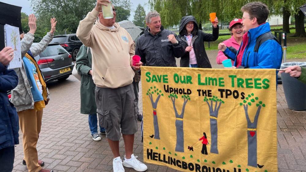 A group of people celebrating, holding drinks in the air with a banner that reads "Save our lime trees"