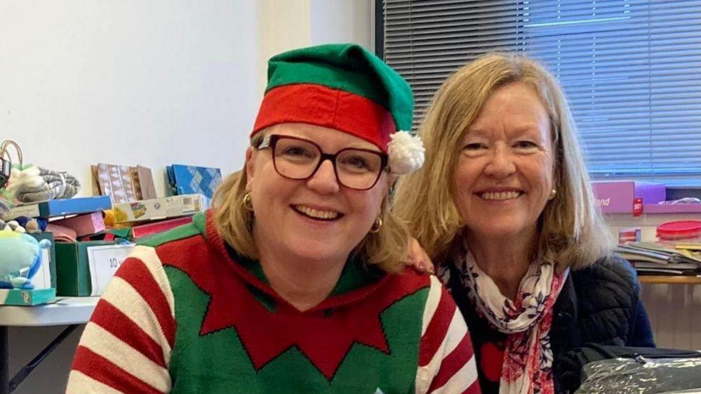 On the left is a woman in glasses wearing a green and red elf hat, with a festive elf-style jumper. On her right is Gill, she's smiling at the camera with medium-long blonde hair. She's wearing a white and red scarf and blue top. They're in an office surrounded by gifts and toys.