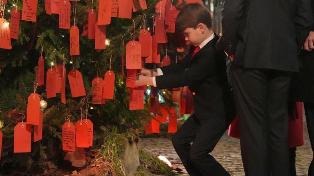 Prince Louis, dressed in a suit, leans down to read messages written on the back of a series of red paper labels tied to a large Christmas tree.