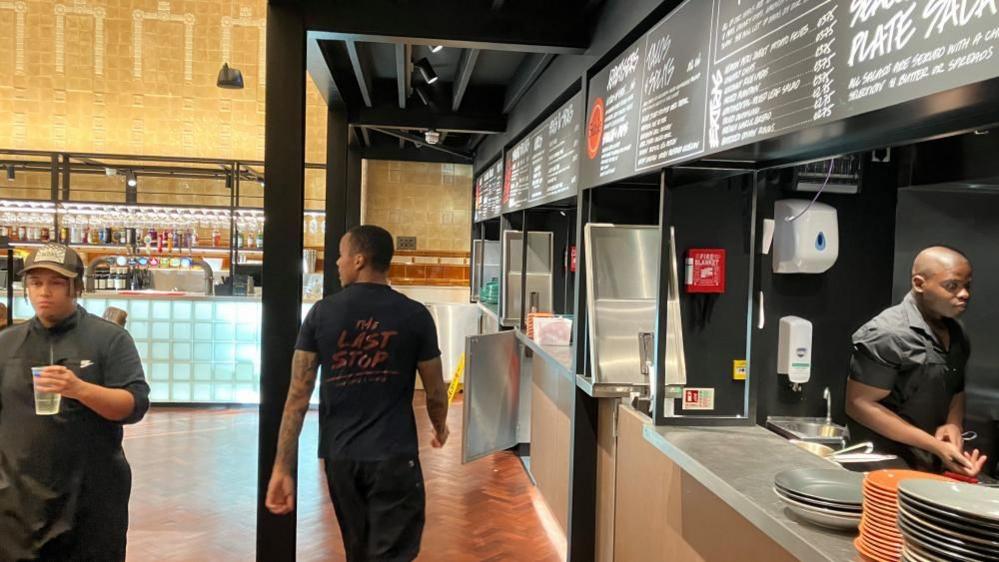 Two staff members are walking in opposite directions in a food hall. A man can also be seen standing behind a bar in an industrial kitchen. 