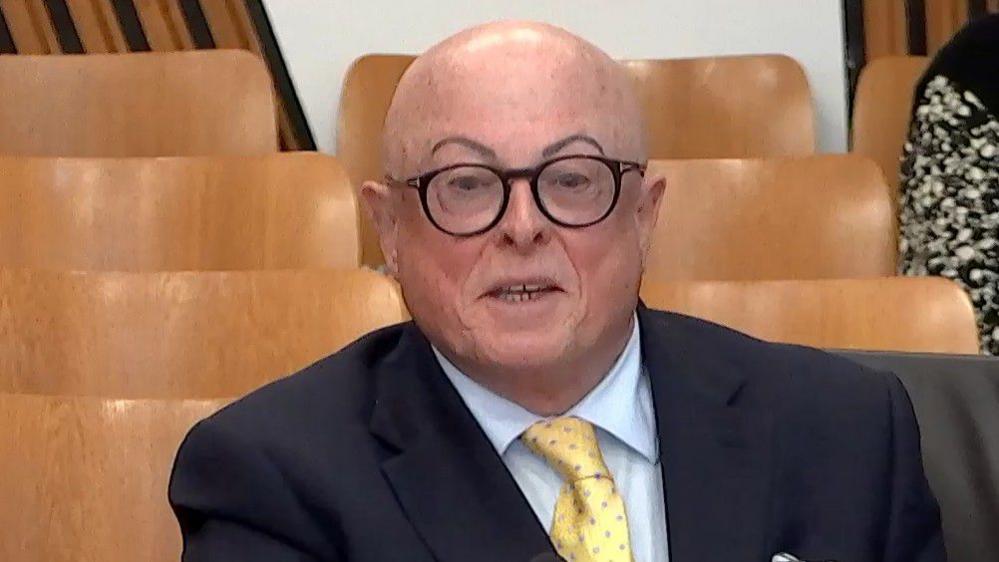 A bald man with glasses, wearing a dark suit and yellow tie