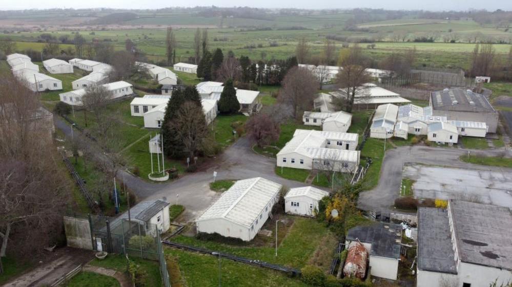 An aerial view of the Northeye prison site. Around a dozen white single-storey buildings are seen with open fields behind them. 