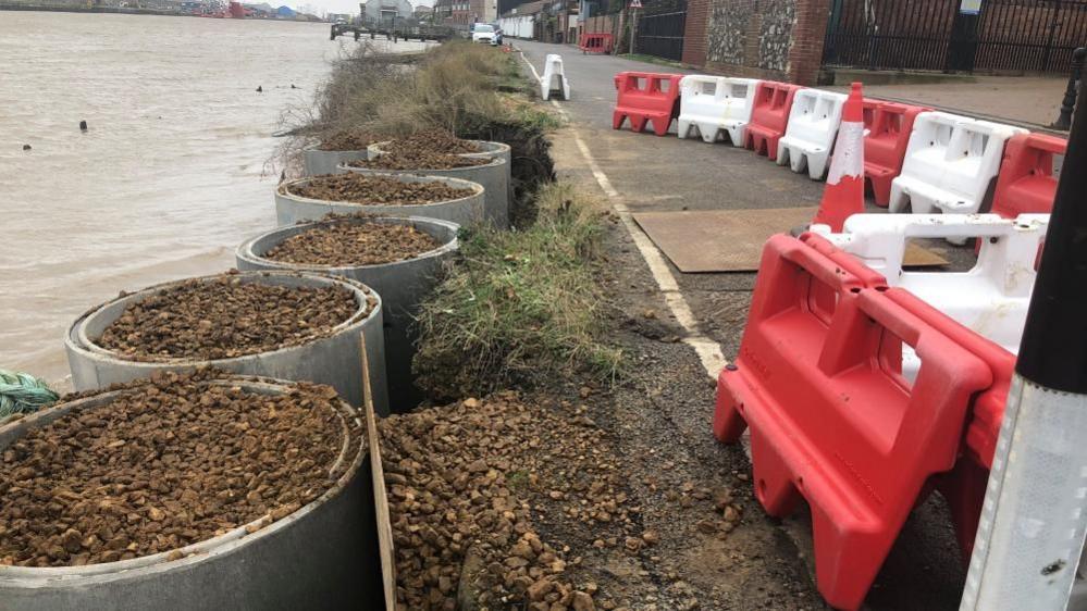Several concrete pipe sections, stood vertically in the river, filled with ballast form a new defence against the bank of Riverside Road. Red and white traffic barriers are in place around a hole in the road that opened up as a result of undermining by river water causing erosion.