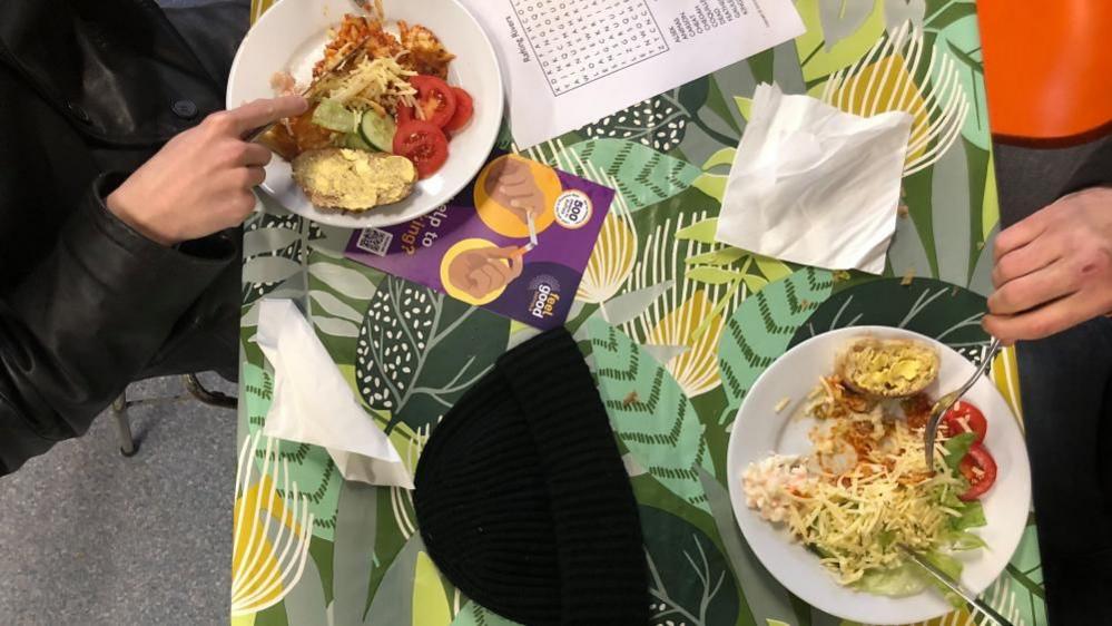 The picture is taken above a table with a brightly covered table cloth. There are two plates full of food, which include baked potatoes, beans, cheese and salad. 