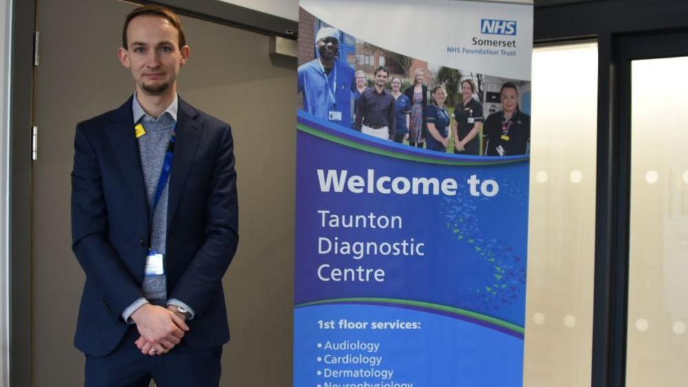 Programme Director David Craig wearing a navy coloured suit at the entrance to the Taunton Diagnostic Centre. There is a poster advertising the services from audiology, cardiology and Neurology. 