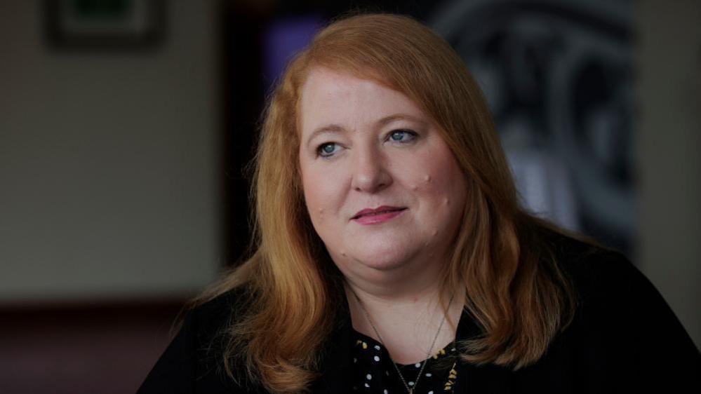 Naomi Long has shoulder length ginger hair and is wearing a black top. she has pink lipstick on. 