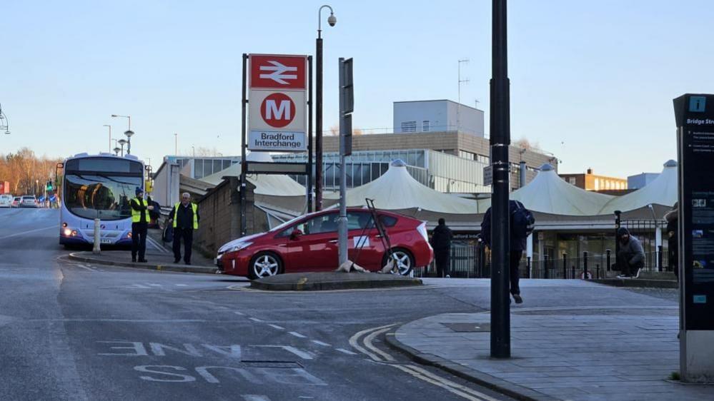 A red car drives out of Bradford Interchange, with two uniformed personnel standing beside a bus on the road. The interchange building with white canopy structures is visible in the background, along with signage for rail and metro services. A few pedestrians are walking nearby.
