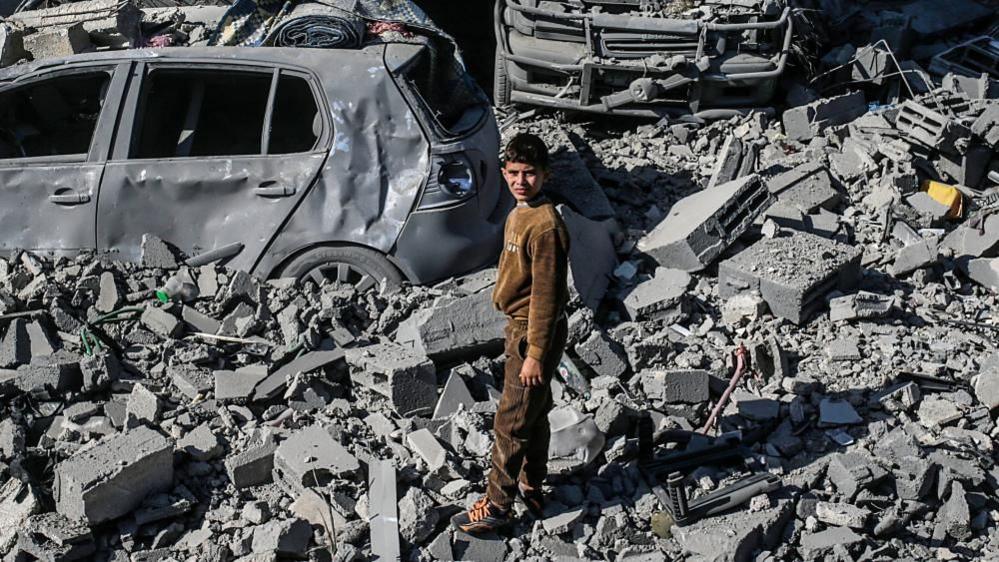 A pre-teen boy stands amongst grey rubble and burned out cars, squinting up towards the camera