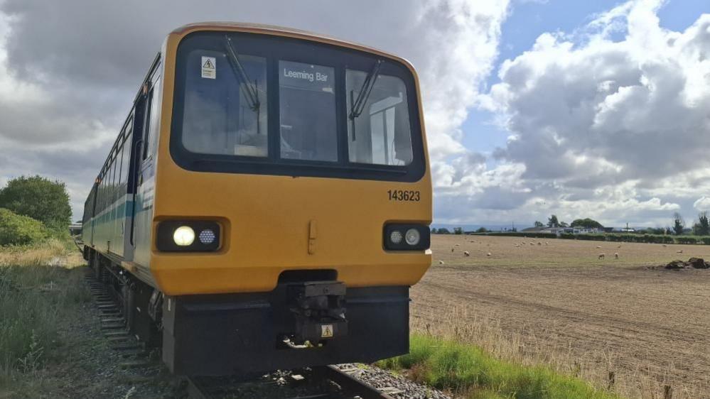 A pacer-type train in the foreground, with sheep in a field in the background. The train has "Leeming Bar" on the destination display.