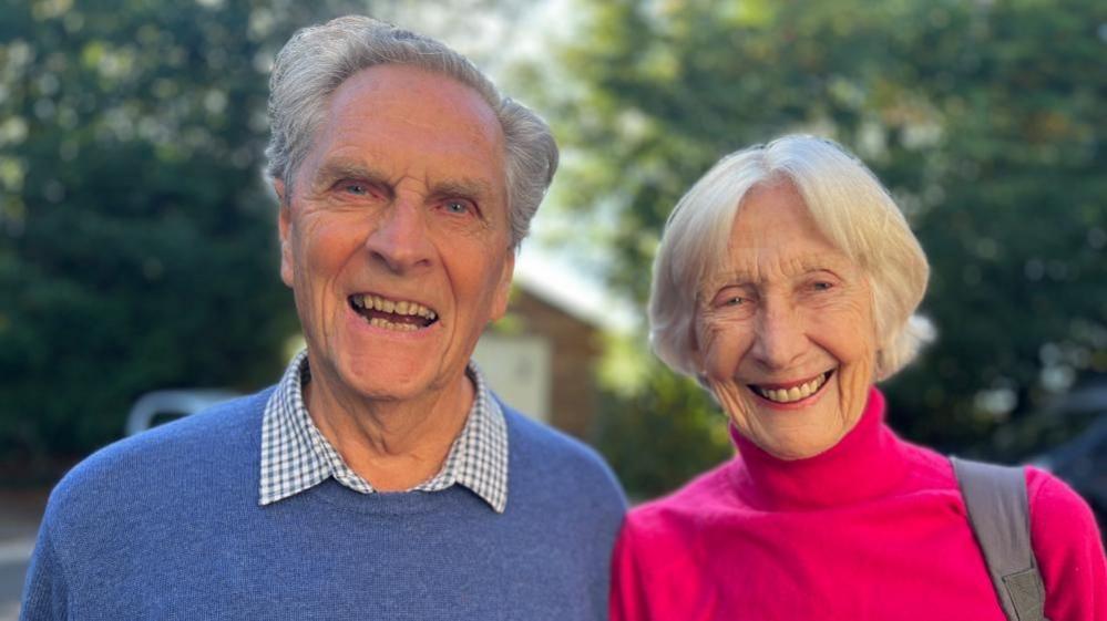 A man in a blue jumper over a checked shirt next to a woman in a bright pink polo neck jumper with a grey bag strap over shoulder smile to camera. 