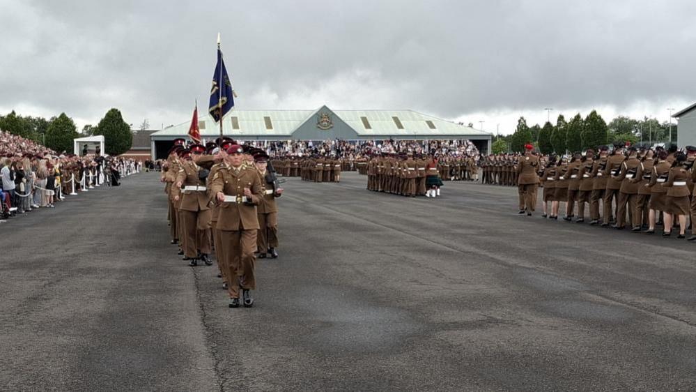 A 16-year-old young man is at the front of a parade dressed in brown military uniform. Hundreds of fellow junior soldiers march behind him, with some carrying flags, and thousands of people watching in the audience.