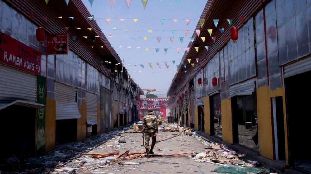A man dressed in military fatigues walks down an empty market, where all the shops have been damaged and broken into. There is debris all over the road.