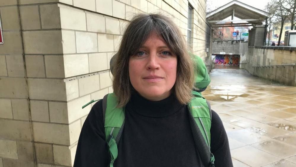 Bethany Mogie with long dark hair, wearing a dark top and carrying a green ruck sack, stands outside a stone-built court building