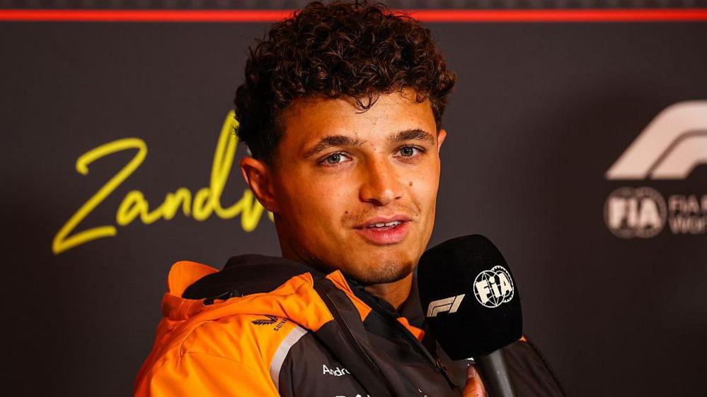 Lando Norris speaking into an F1 and FIA-branded microphone at a news conference before the Dutch Grand Prix. He is wearing a McLaren rain jacket