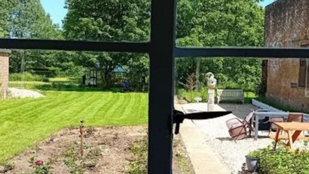 A view through a 1930s Crittall window, looking out onto the grounds of the former RAF West Raynham headquarters building. It shows laws to the left, and a garden set with shrubs, and a stone area where seating and tables and a sculpture have been placed. There are trees in the backdrop, and a red brick wall to the right of the image.