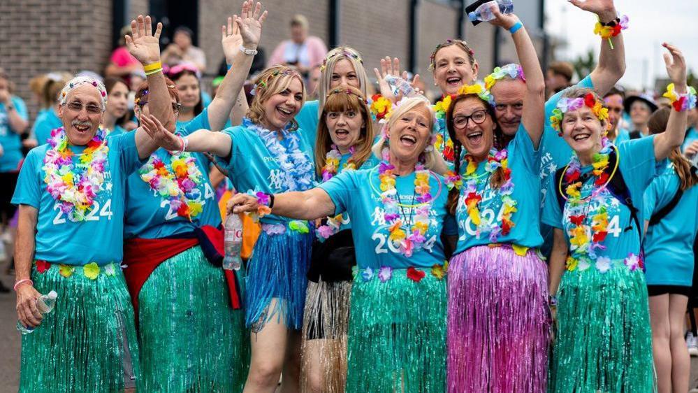 A group of people wearing bright fake flowers and skirts, looking at the camera and cheering 