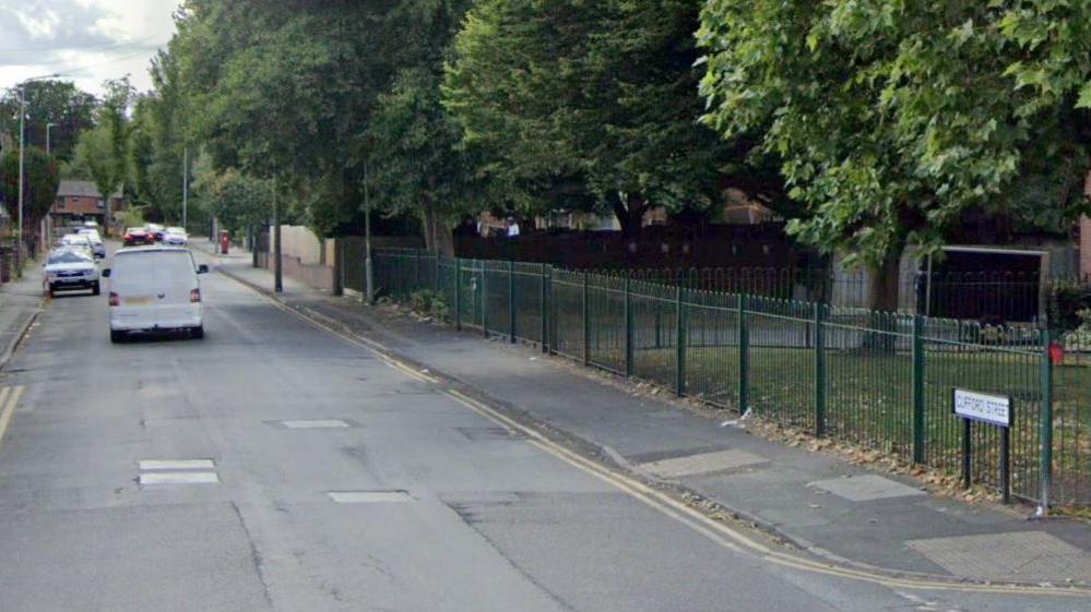 A white van drives on Clifford Street in Wolverhampton. Double yellow lines can be seen on the side of the road, alongside the street sign. Green railings and grass can be seen to the right of the image. Other cars can be seen in the background.