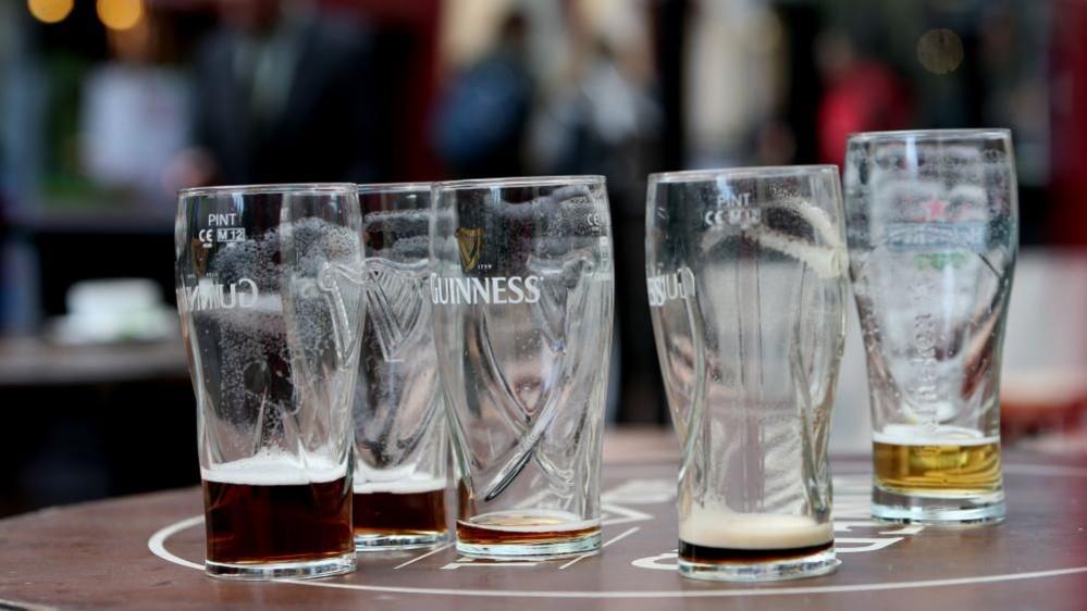 Five empty beer glasses on a brown table. Four of these are Guinness glasses, with small amounts of beer in the bottom, and the one on the furthest right is a Heineken glass, with a small amount left.