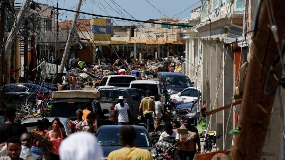 A crowded street filled with people and vehicles navigating wreckage - collapsed power lines hang low over the road and debris from damaged buildings is scattered everywhere