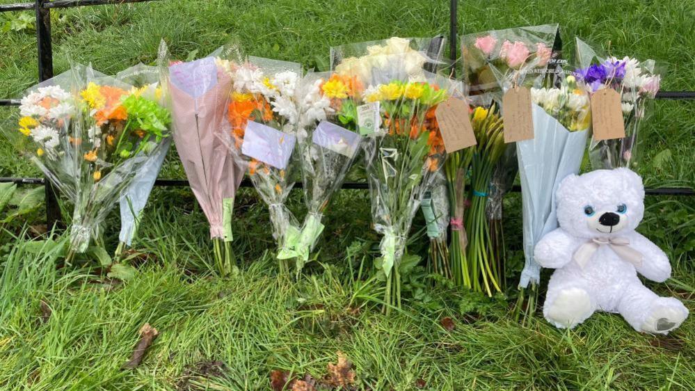 Bouquets of flowers, some with handwritten notes are propped against metal railings along with a single white teddy bear