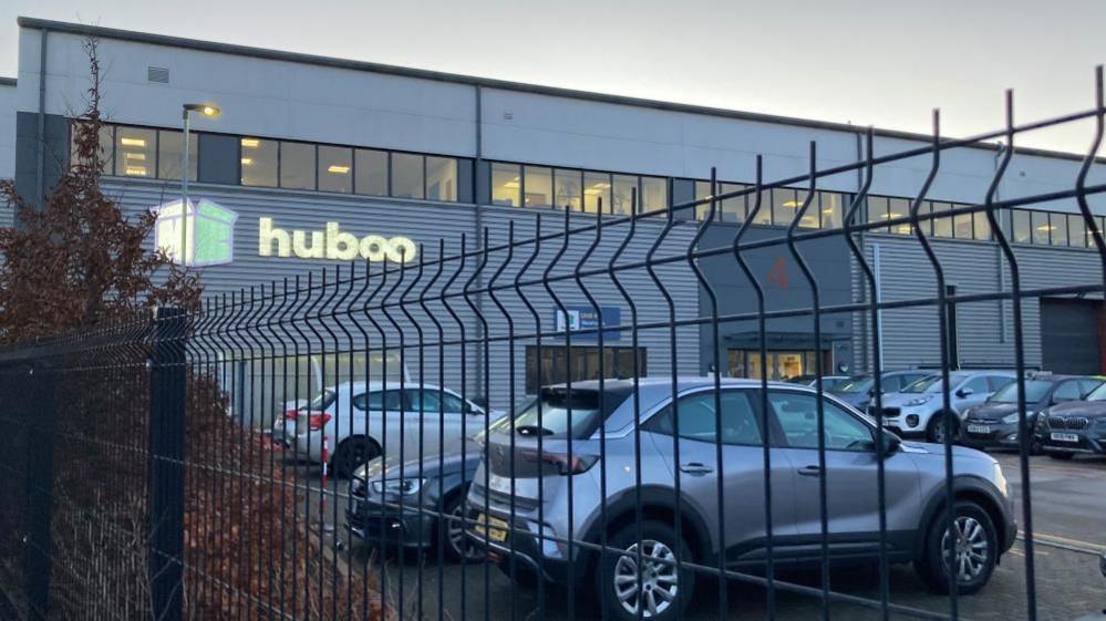 An exterior view of Huboo's warehouse site, with cars and a fence in the foreground.