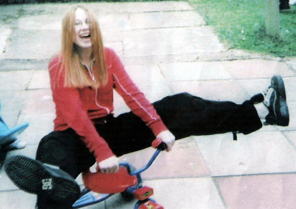 A young girl, possibly a teenager, sits on a children's bike with her legs kicked out in the air on either side. She wears black baggy trousers, black shoes, and a red zipped up hoodie. She also wears glasses and has long straight light brown hair.
