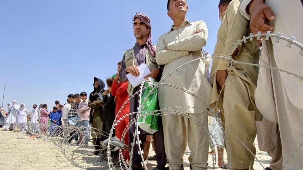 Afghans wait patiently in line in Kabul in 2021 to flee on evacuation flights, some with their arms crossed, with barbed wire in the foreground.