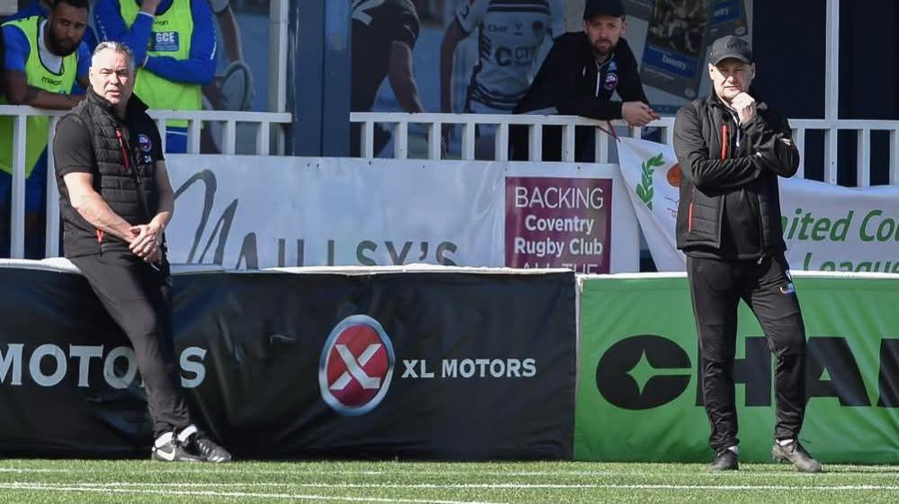 Darren Acton sits on an advertising board watching on with joint-manager Russell Dodd on the touchline during a match