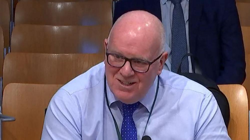 A bespectacled man in a light blue shirt and blue tie