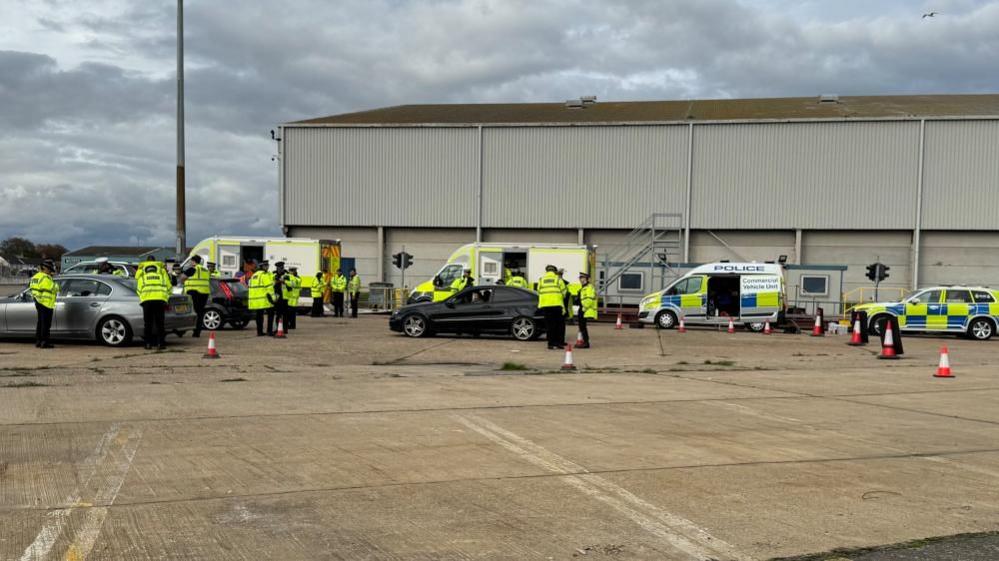 A line of police and other agency vehicles lined up on hard standing near a portside warehouse, with other vehicles being checked by police.