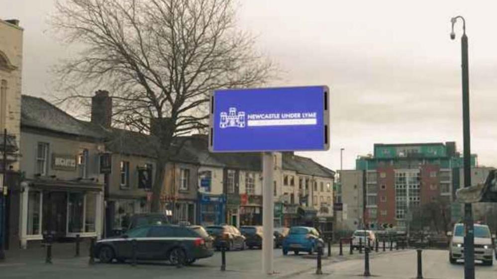 Computer generated image of proposed giant screen in Newcastle under Lyme town centre alongside buildings and parked cars.