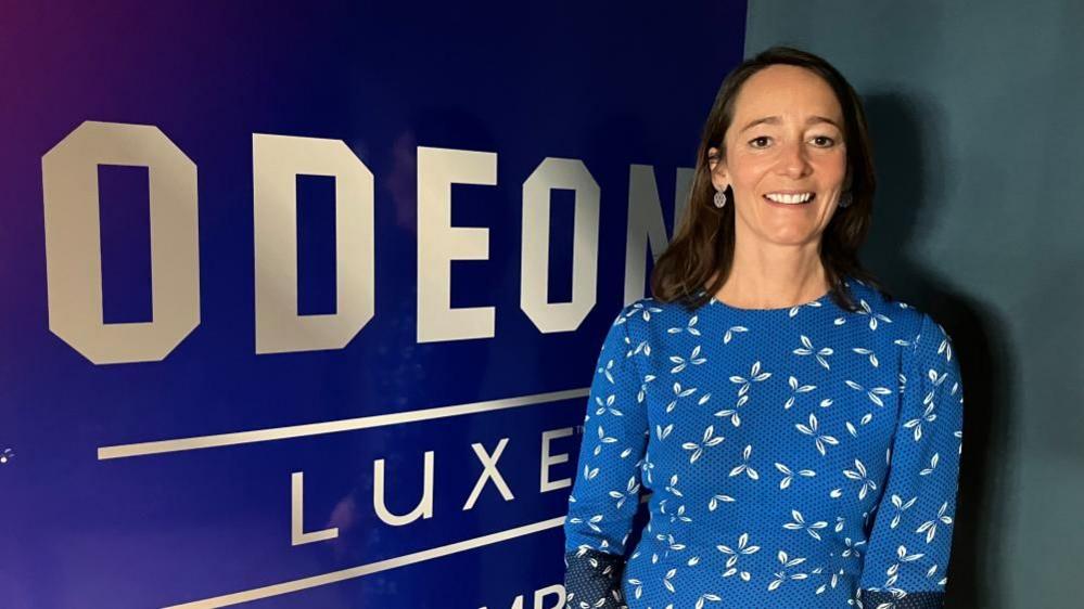 A woman in a blue dress, smiling and looking at the camera, next to a Odeon LUXE sign.