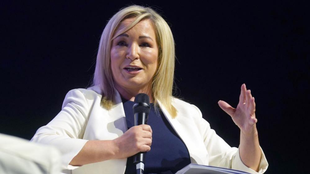 Blonde woman in white blazer speaking into a microphone 