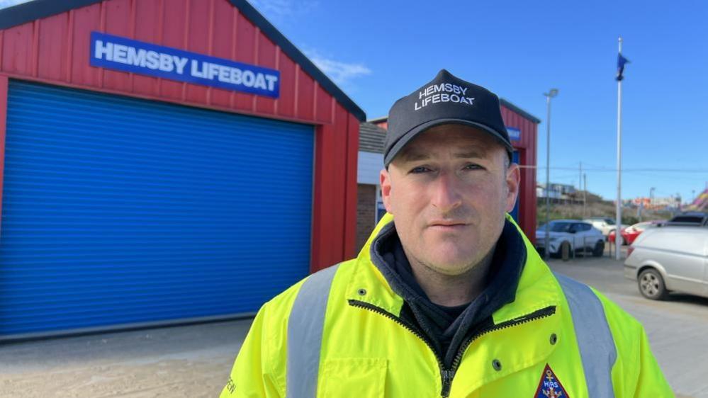 Dan Hurd, wearing a black baseball cap and a yellow hi-vis coat, both branded "Hemsby Lifeboat". He is standing in front of the current lifeboat station, which is a red-clad building with blue roller-shutter doors.