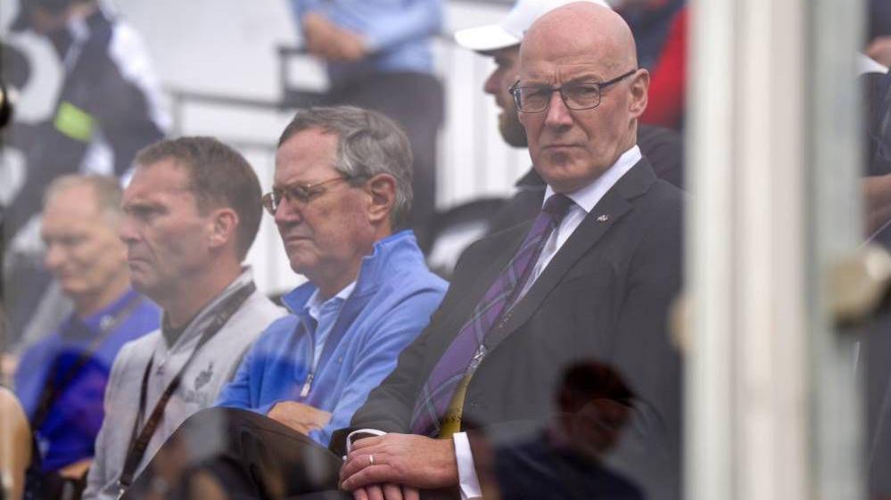 John Swinney, a bald man with glasses, wearing a dark suit and purple tartan tie. He has a stern expression and appears to behind some sort of glass screen. 
