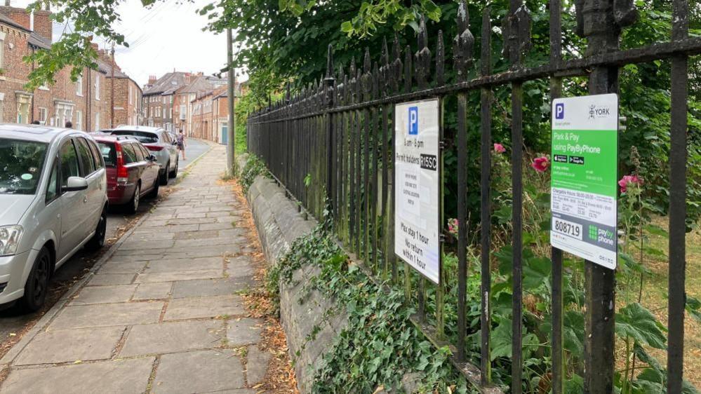 A view of Pay By Phone parking signs, affixed to black metal railings on Bishophill Senior in York. Cars are parked by the side of the road, and red brick houses can be seen in the distance.