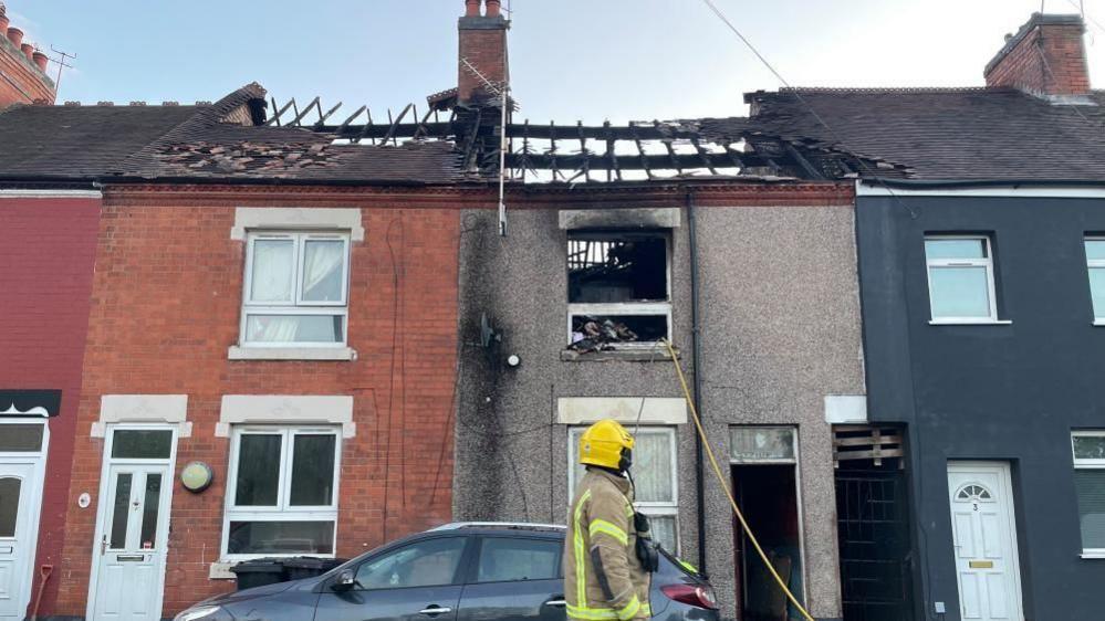 A residential street where a fire has ripped through the roof of three properties. A firefighter is in front of the houses and a grey car is also parked there.