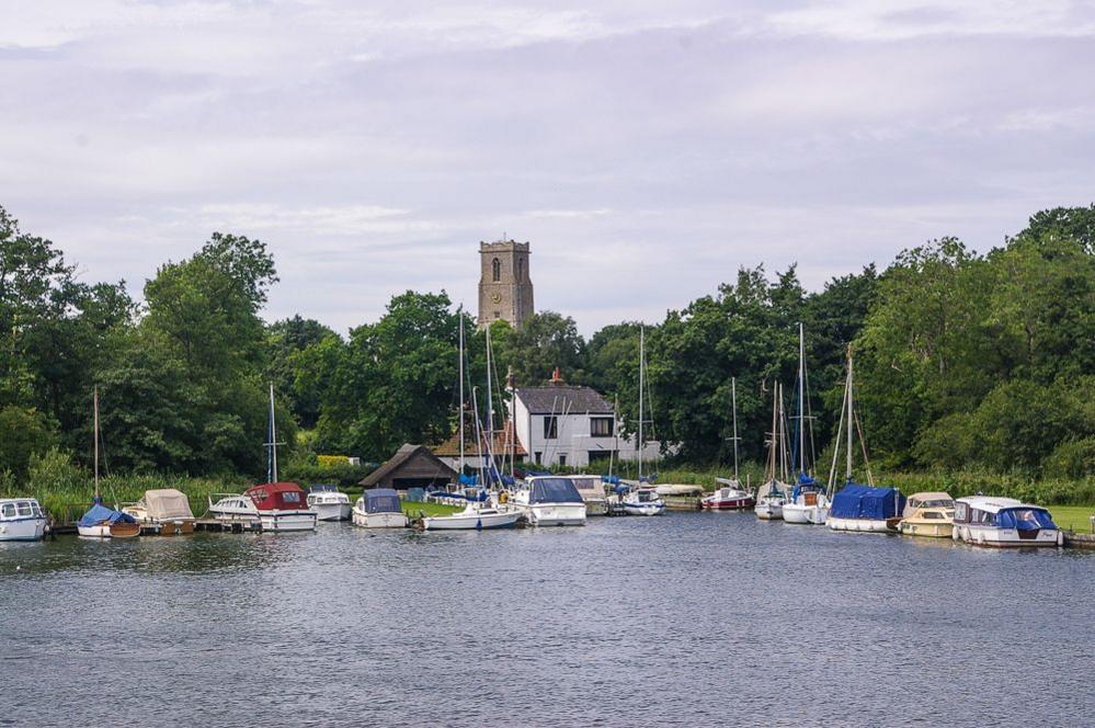 Church tower in distance. In foreground is an expanse of river water with about 20 boats of different styles, including sailing boats and mini cruisers moored outside a building which looks like a pub. 