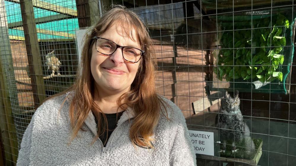 Owl sanctuary founder Louise Thorpe, next to a cage with an owl sitting on a shelf. Louise is wearing a grey fleece top, and a black teeshirt underneath. She is wearing glasses, has long auburn hair, and she is smiling.