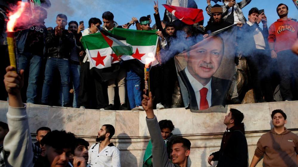 A crowd celebrating with flares and holding up flags of Syria and one of President Erdogan