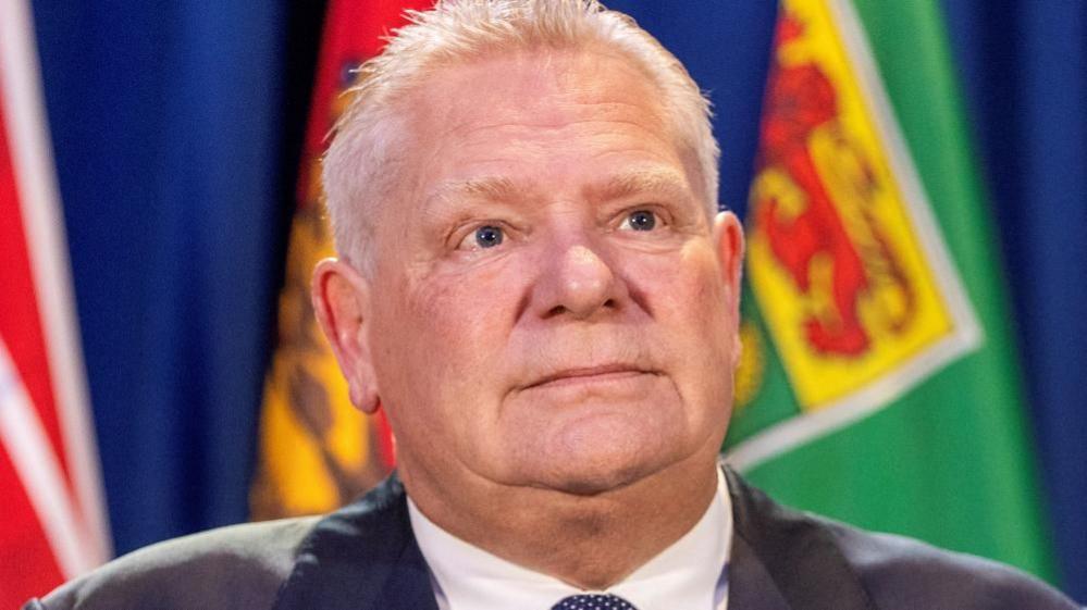 A close-up profile image Ontario Premier Doug Ford with provincial flags hanging in the background. 