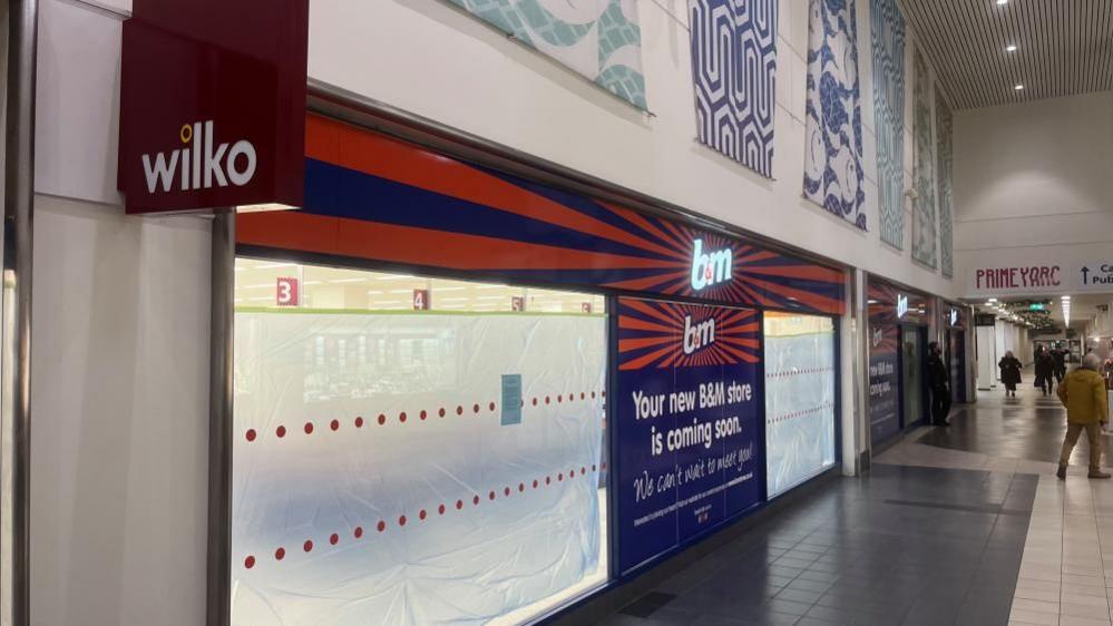 Wilko branded sign adorns the shopping centre facade with new B&M Bargains signage now showing, with polythene screens in the shop window to obscure refitting work inside. To the right of the image is the shopping centre, featuring grey and white tiled floor, and abstract design panels above the shop facade.