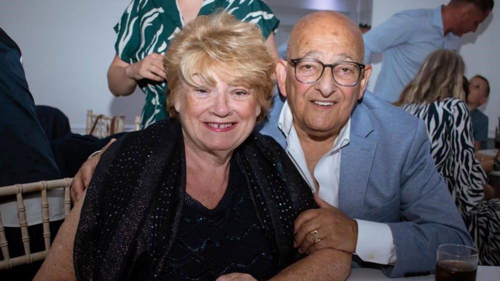 Elaine has short blonde hair and wears a navy blue dress with a scarf around her shoulders. Philip has his arm around her and is wearing a light grey blazer and white shirt