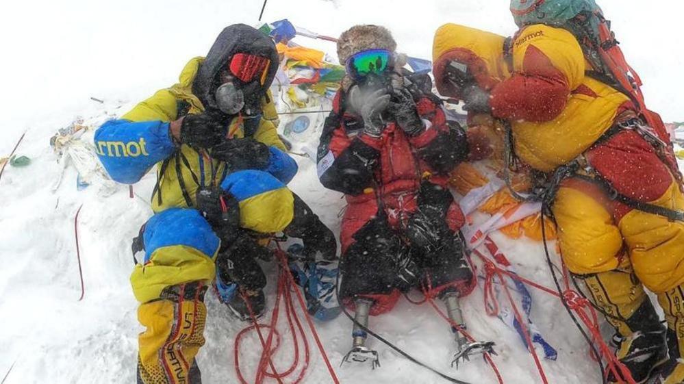 Hari Budha Magar MBE, with two climbers at the top of Mount Everest. He is wearing a red coat with both of the other climbers wearing yellow coats. They are on top of a snow covered mountain peak.